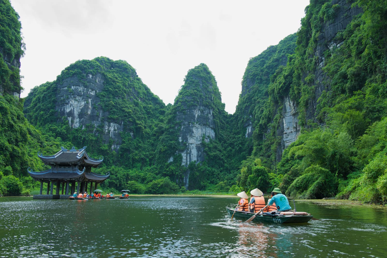 Rowboats of visitors in life jackets gliding past a pagoda beneath limestone cliffs in Trang An